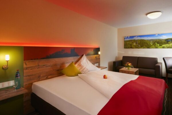 Themenbild von einem Hotelbett in einem modernen Zimmer mit brauner Ledercouch und roter Hintergrundbeleuchtung am Bett