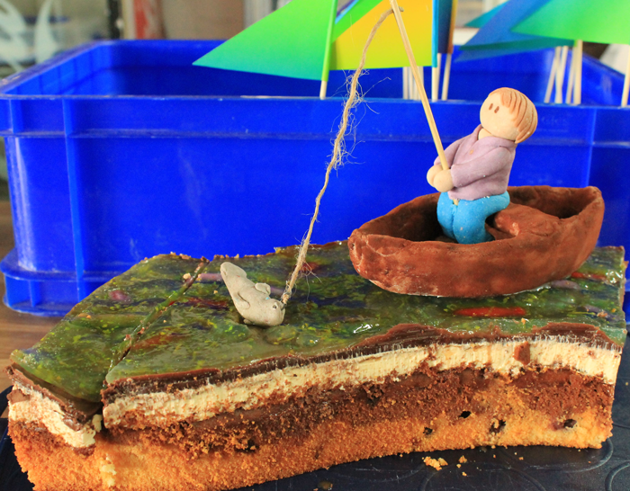 Bild von einem Stück Donauwelle mit modelliertem Boot mit Insasse, der einen Fisch an der Angel hat vor einer blauen Kunststoffkiste