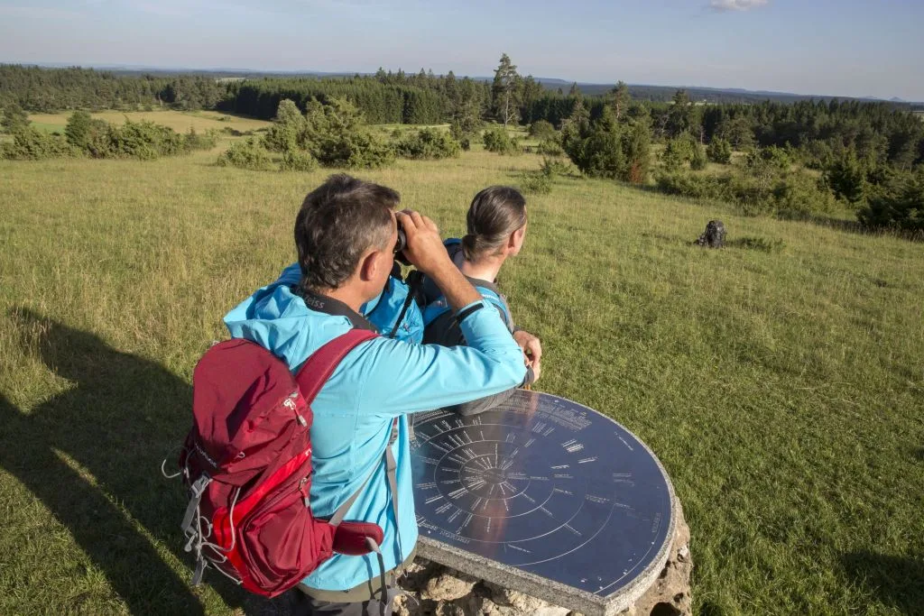 Bild von zwei Wanderern, die vom Alten Berg aus die Fernsicht genießen. Sie stehen auf einer Wiese an einer Landschaftsübersichtskarte
