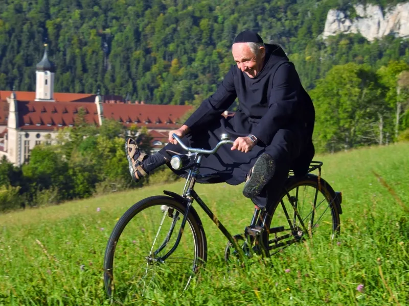 Picture of a monk riding a bicycle on a meadow and stretching his legs. A monastery can be seen in the background