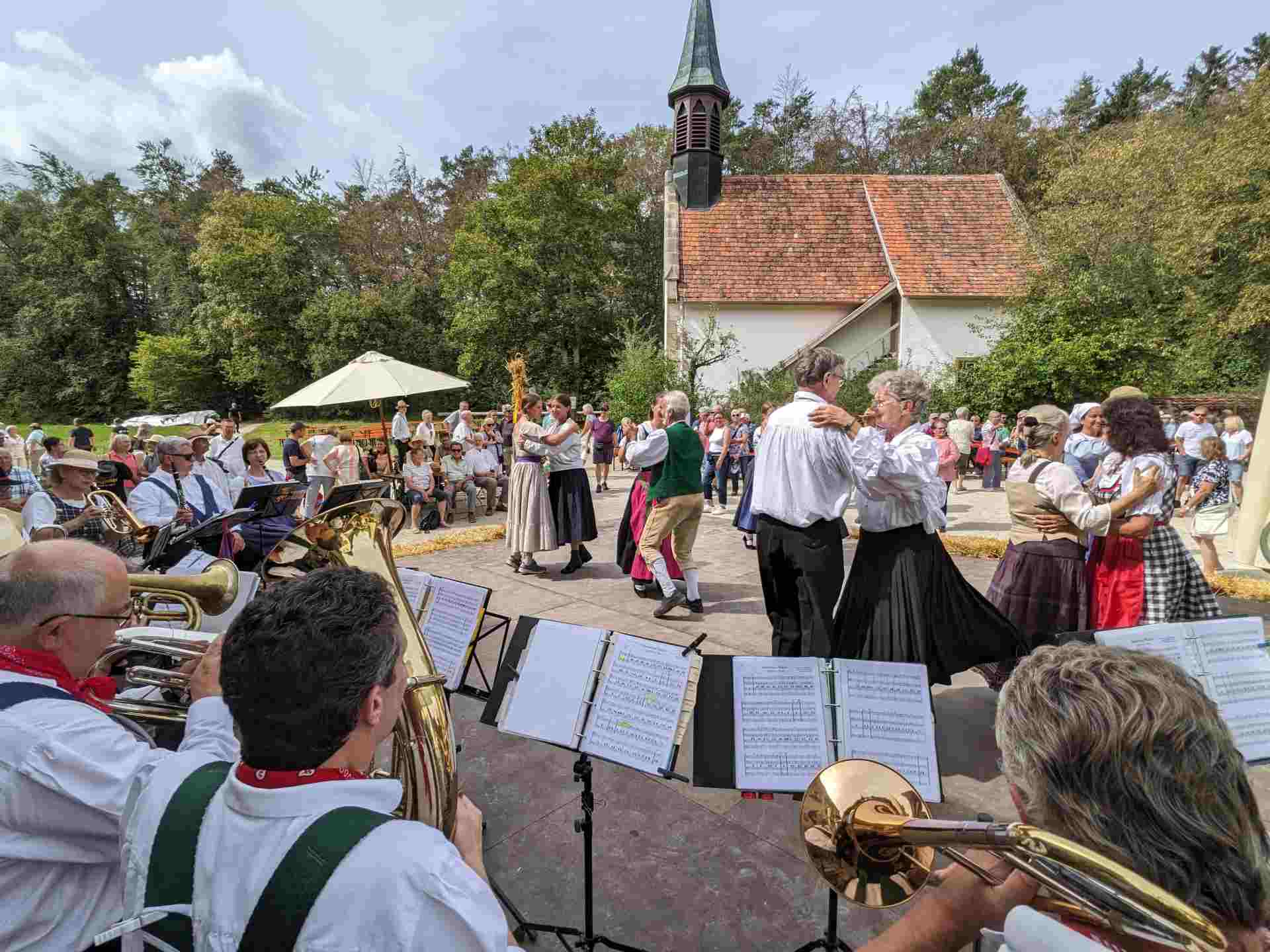 Tanzende Paare in Tracht unter freiem Himmel auf einer Bühen auf dem Dorfplatz mit Musikkapelle