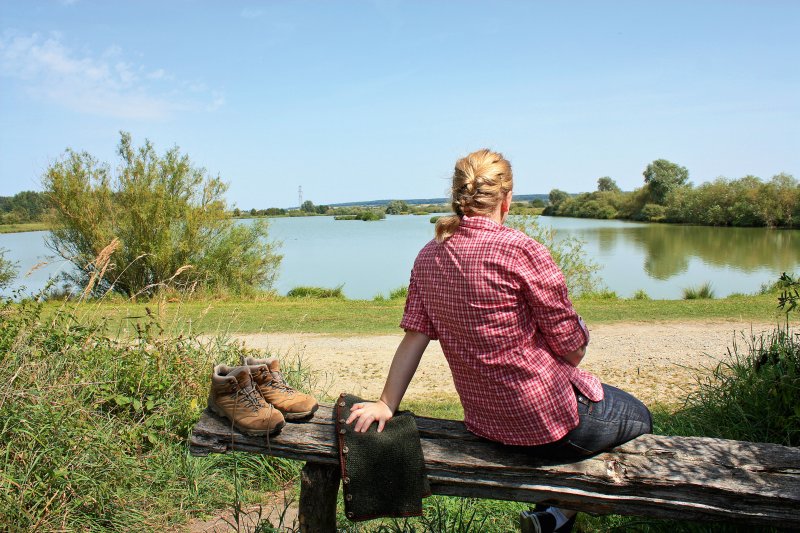 Frau auf einer Bank am See blickt mit dem Rücken zum Fotografen auf den See, hat Wanderschuhe ausgezogen