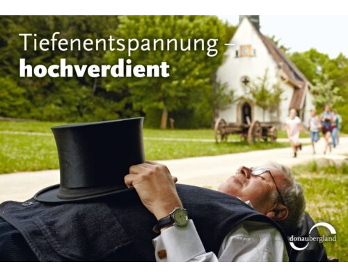 Donaubergland Postkartenmotiv mit schlafendem Mann mit Zylinder auf dem Bauch vor einer Kapelle.